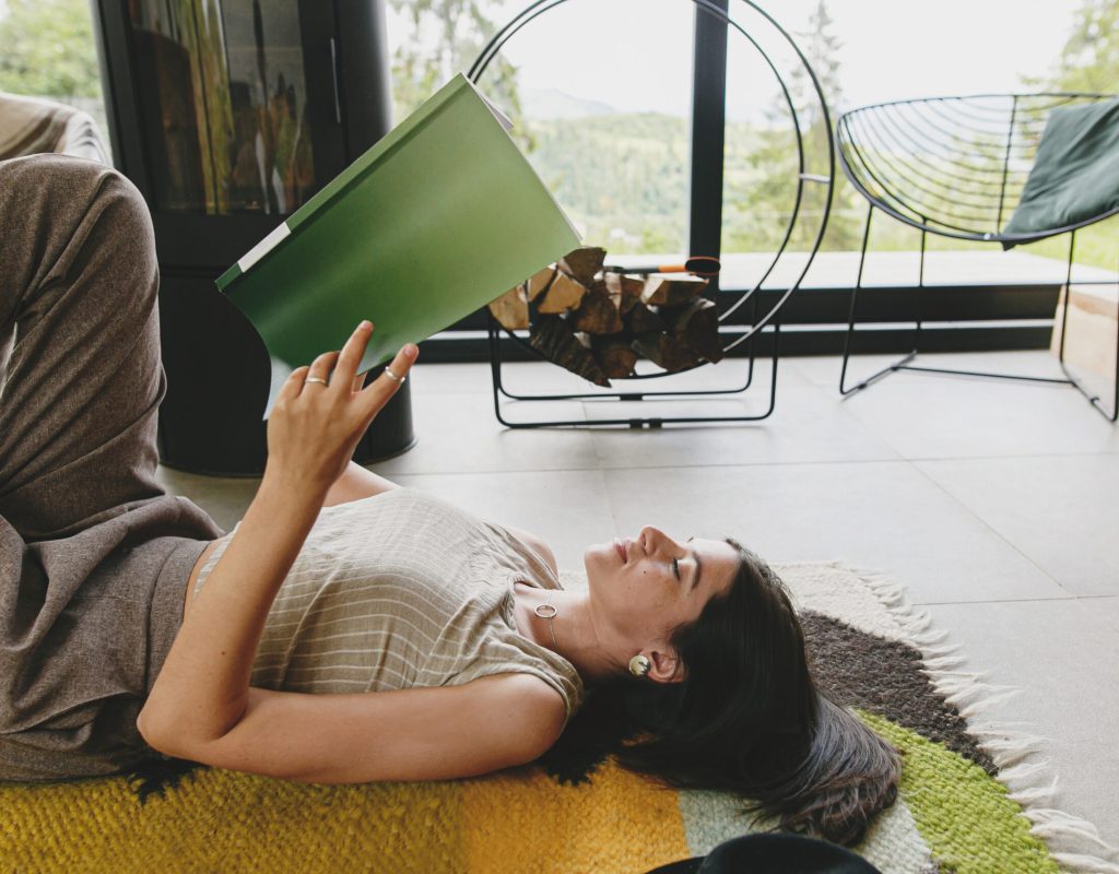 Beautiful stylish woman relaxing with book on cozy rug on background of fireplace in modern chalet. Young female in casual clothes reading magazine on floor. Leisure and vacation time