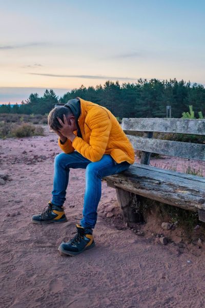 A sad older male clutching his head in his hands on an old wooden bench on a rural field